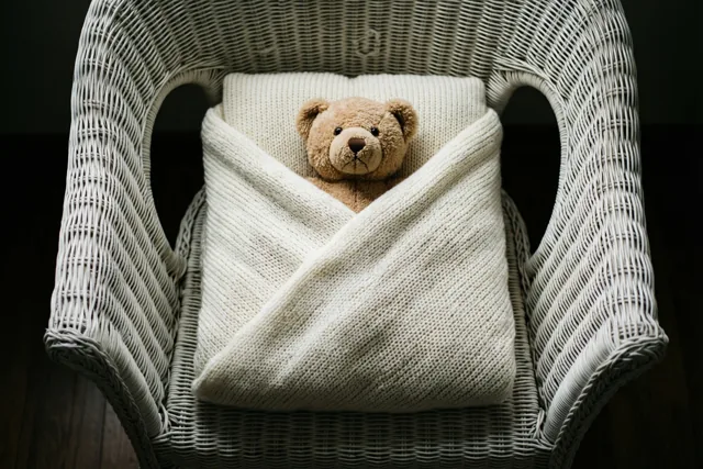 Een kleine bruine teddybeer zit gewikkeld in een crèmekleurige gebreide deken op een witte rieten stoel tegen een donkere achtergrond.