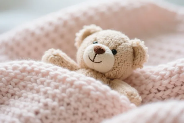 Een klein gebreid teddybeertje ligt in een zachte, pastelroze gebreide deken bij gedempt licht.