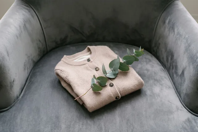 Een opgevouwen beige babyvestje ligt met een takje eucalyptus op een grijze fluwelen fauteuil in zacht licht.