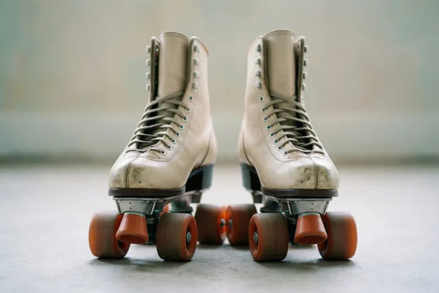 Een paar oude rolschaatsen met beige leren schoenen en oranje wielen staat naast elkaar op een lichtgrijze vloer.