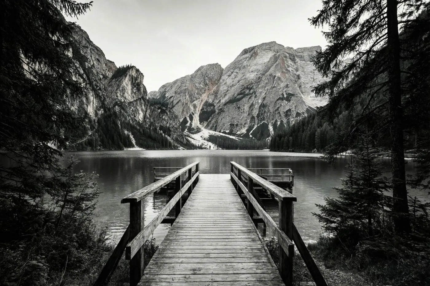 Een zwart-witfoto toont een houten steiger op een kalm bergmeer, met dennen aan weerszijden en hoge rotsbergen op de achtergrond.