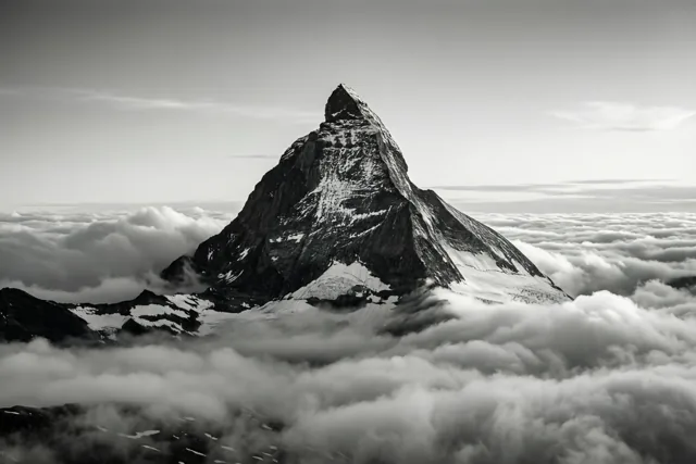 Een scherpe bergtop met een laagje sneeuw steekt boven een dicht wolkendek uit in een zwart-witbeeld.