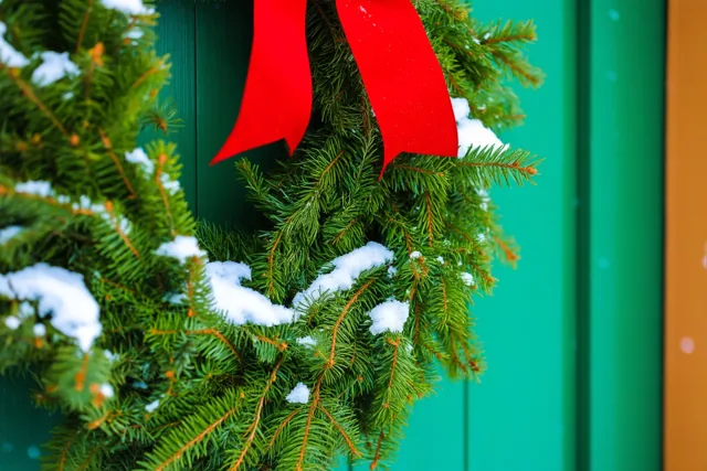 Een groene kerstkrans met sneeuw en een rood lint hangt aan een groene houten deur en is van dichtbij in beeld gebracht.
