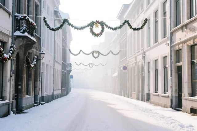Een besneeuwde straat in Leuven wordt omzoomd door grijze gebouwen met groene kerstslingers en kransen.