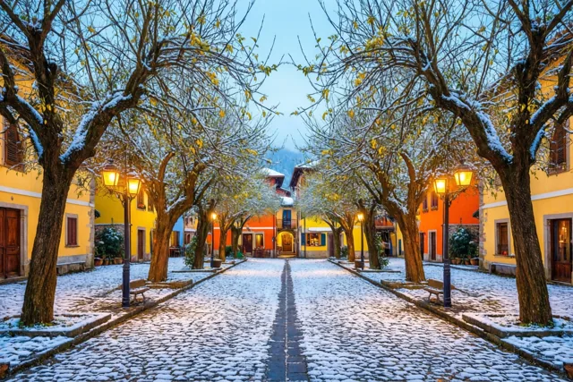 Op een besneeuwd dorpsplein met kasseien staan kale lindebomen met sneeuw, brandende straatlantaarns en kleurrijke gebouwen onder een schemerhemel.