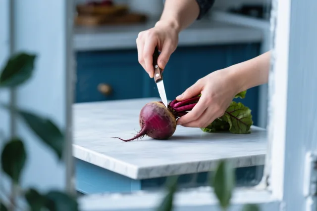 Een persoon snijdt een suikerbiet op een wit marmeren keukenblad, met blauwe kasten en groene planten op de achtergrond.