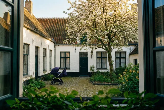 Door een raamkozijn is een rustig binnenplein met witte huizen te zien, met een kinderwagen bij een deur onder een bloeiende boom in zacht warm licht.