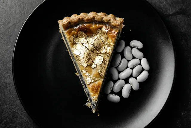 Een stuk galette met bladgoud ligt op een zwart bord naast een hoopje witte bonen tegen een donkere achtergrond met textuur.