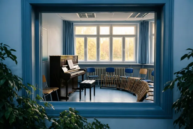 Door een blauw raamkozijn is een muzieklokaal te zien met een staande piano, blokfluiten in een boog en stoelen in een halve cirkel, met blauwe muren en gordijnen in warm zonlicht.
