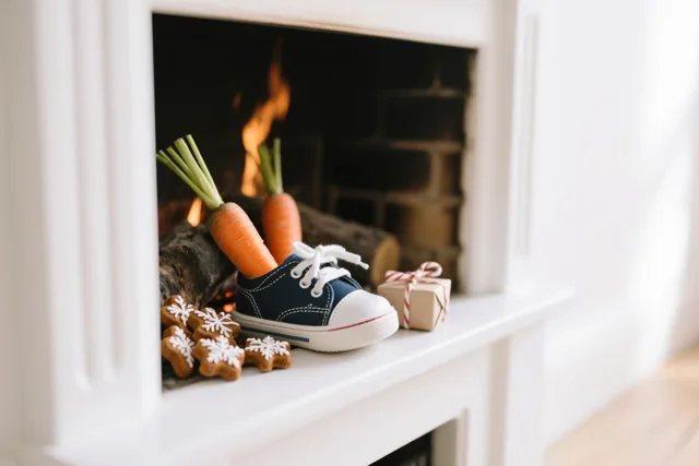 Een kleine blauwe kindersportschoen staat op een witte schoorsteenmantel naast sneeuwvlokvormige speculaas en een ingepakt cadeau, met wortels en vuur in de haard.
