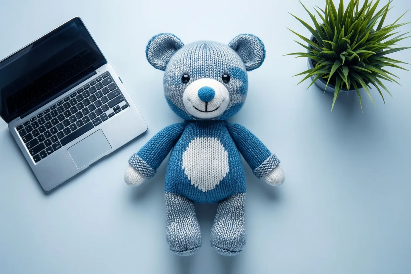 Een blauw-witte gebreide teddybeer zit op een lichtblauw oppervlak naast een kleine zilverkleurige laptop en een groene plant in een pot.