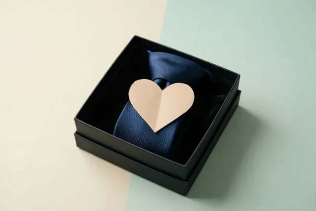 Een donkerblauwe zijden das ligt in een zwarte geschenkdoos met een lichtbeige papieren hart erop, tegen een beige en mintgroene achtergrond.