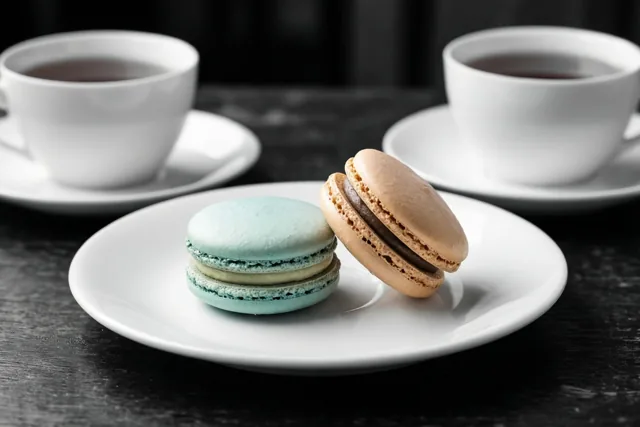 Een lichtblauwe en een beige macaron liggen op een wit bord tussen twee witte theekopjes op een donkerhouten oppervlak.
