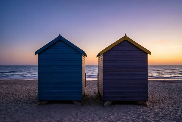 Een blauwe en een paarse houten strandcabine met gele afwerking staan naast elkaar op het zand bij zonsondergang, onder een hemel die van paars naar oranje overgaat.