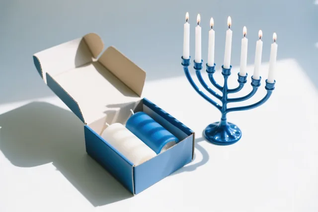 Een open blauwe kartonnen doos met blauwe en witte kaarsen staat naast een blauwe kandelaar met brandende witte kaarsen op een wit oppervlak met sterke schaduwen.