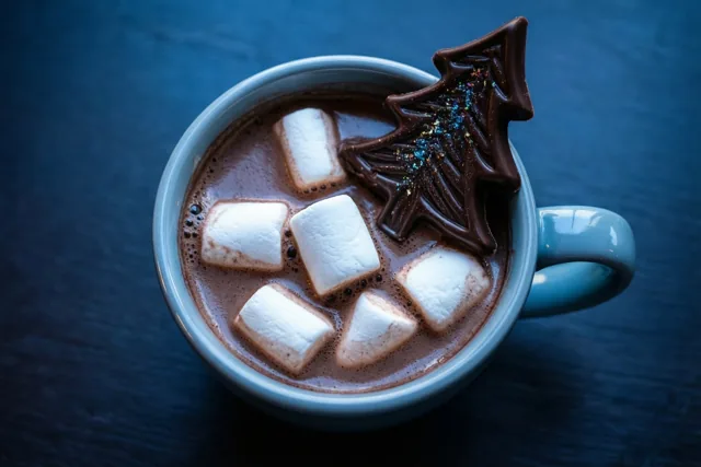 Van bovenaf gezien staat een lichtblauwe mok warme chocolademelk met witte marshmallows en een roerstaafje in de vorm van een chocolade kerstboom op een donkerblauwe ondergrond met reliëf.