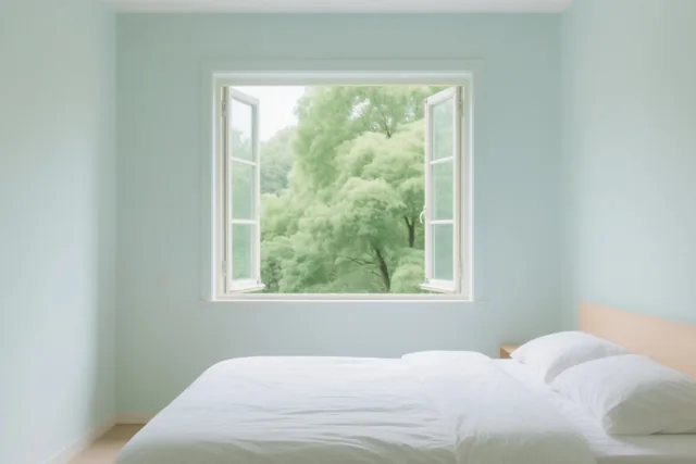 Een lichte slaapkamer heeft lichtblauwe muren, een bed met wit beddengoed en een open raam met zicht op groene bomen.