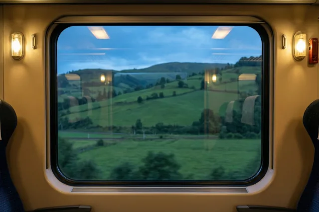 Warm licht valt op beige wanden en donkerblauwe treinzetels, met door het raam groene heuvels onder een blauwe hemel.