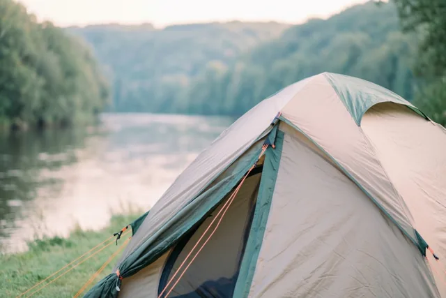 Een beige en blauwgroene tent staat in zacht licht op een grasrijke oever bij een kalme rivier en beboste heuvels.