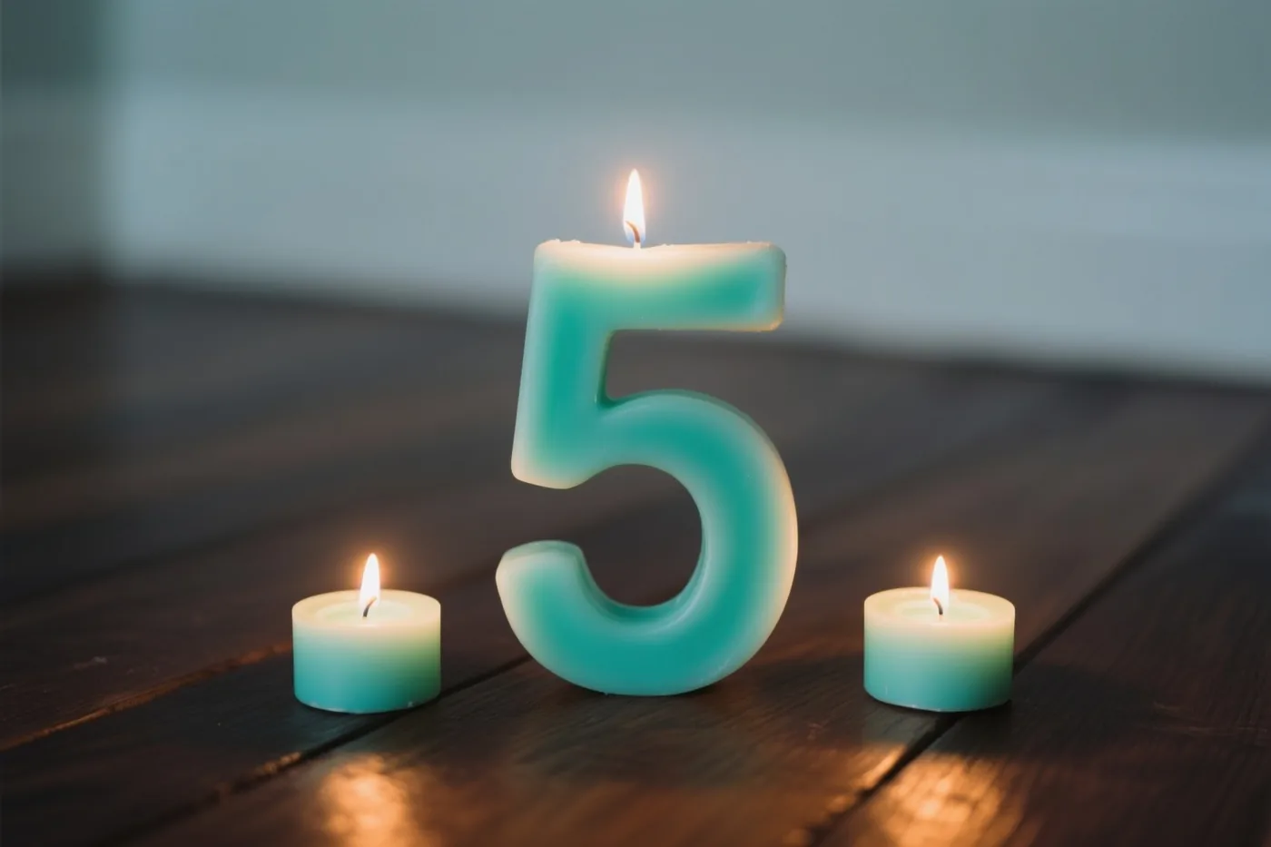 Een grote blauwgroene kaars in de vorm van een 5 en twee kleinere blauwgroene kaarsen branden op een donkere houten vloer tegen een donkere achtergrond.