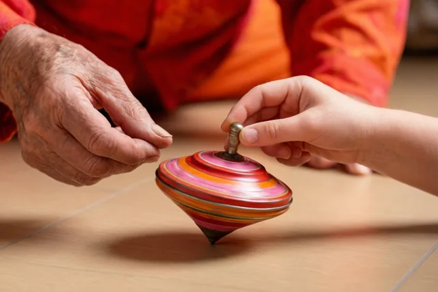 Een oudere hand en een kinderhand laten samen een kleurrijke blikken tol draaien op een lichte houten vloer, met rode, oranje en roze tinten.