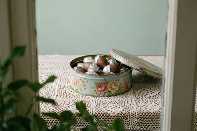 Een open oud bloemenblik met chocoladepaaseitjes in folie staat op een crèmekleurig gehaakt tafelkleed, gezien door een witte omlijsting met groen gebladerte op de voorgrond.