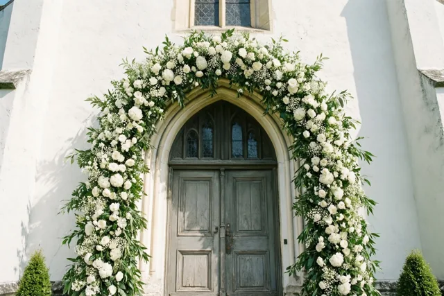 Een volle slinger van witte bloemen en groen hangt in een boog boven een verweerde houten kerkdeur in een witte gepleisterde muur, met een glasraam erboven.