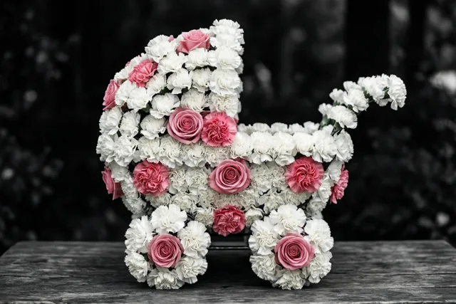 Een bloemstuk in de vorm van een kinderwagen van witte anjers en roze rozen staat op een houten ondergrond tegen een donkere, onscherpe achtergrond.