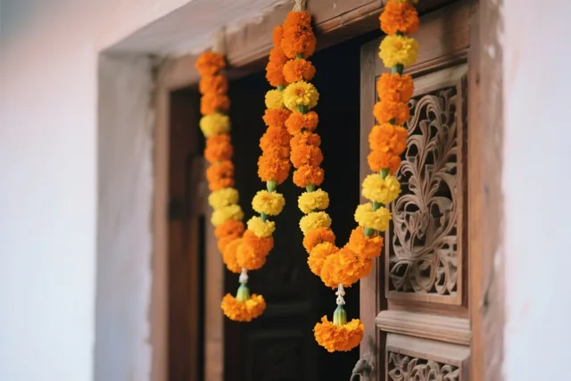 Dikke slingers van oranje en gele afrikaantjes hangen aan een houten deuropening met snijwerk, warm verlicht tegen een donker interieur.