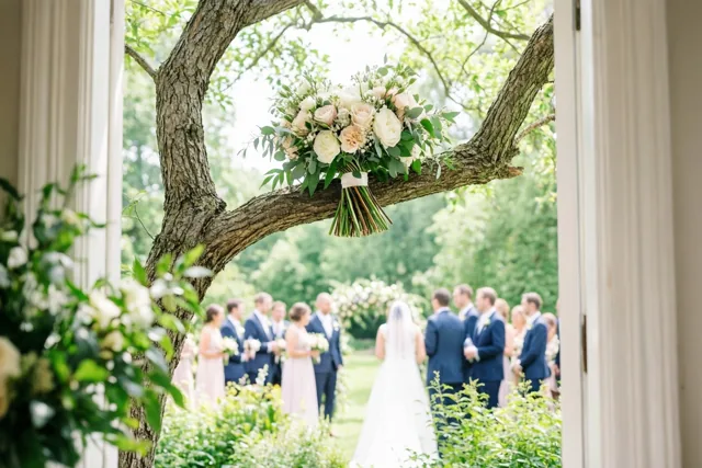 Door een deuropening is een bruidsboeket met witte en lichtroze rozen aan een boomtak te zien, met feestelijk geklede gasten in een groene tuin op de achtergrond.