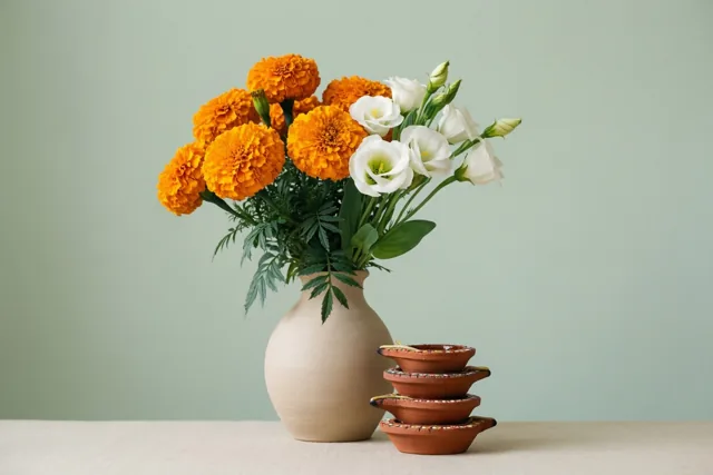 Een vaas met oranje afrikaantjes en witte tuberozen staat naast vier gestapelde terracotta olielampjes op een licht oppervlak, voor een effen bleekgroene muur.