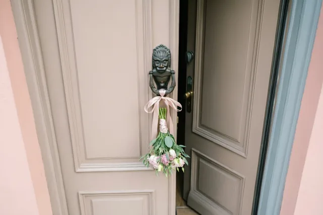 Een boeketje roze en witte bloemen met een lichtroze lint hangt aan een zwarte deurklopper op een lichtbeige voordeur die op een kier staat, met roze en blauwgrijze muren eromheen.