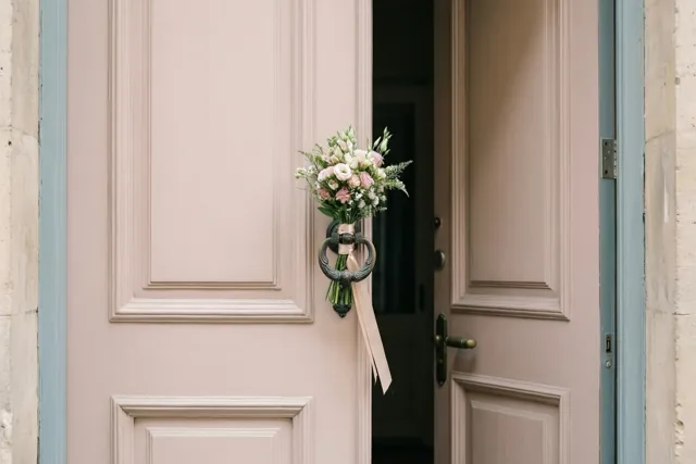 Een boeketje roze en witte bloemen met een lint hangt aan een sierlijke deurklopper op een open lichtroze voordeur, met een lichtblauw deurkader en stenen muren.
