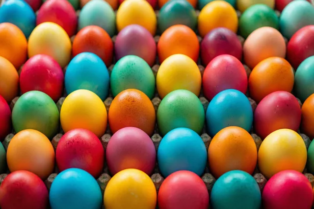 Van bovenaf zijn rijen rood, geel, blauw en groen beschilderde eieren in een kartonnen eierdoos te zien.