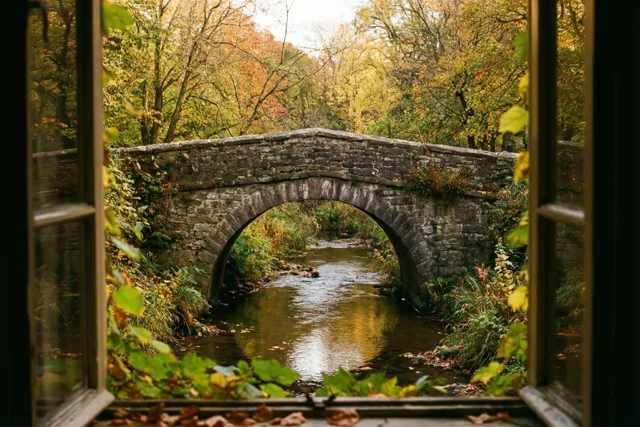 Door een open raam is een oude stenen boogbrug over een rustige beek in een herfstpark te zien, tussen warmgele en groene bladeren.