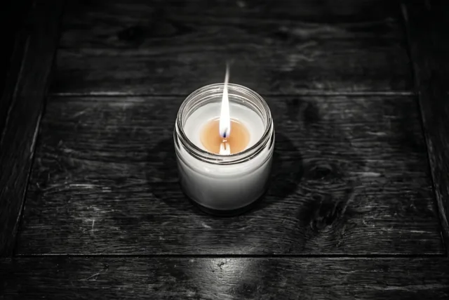 Een kaars in een glazen pot brandt op een donkere houten tafel, met een warm oplichtende vlam in een verder monochroom beeld.