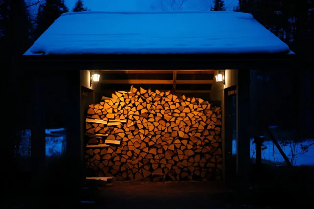 Een stapel gekloofd brandhout ligt onder een besneeuwde houten overkapping, verlicht door oranje lantaarnlicht tegen een donkere nacht en blauwe sneeuw.