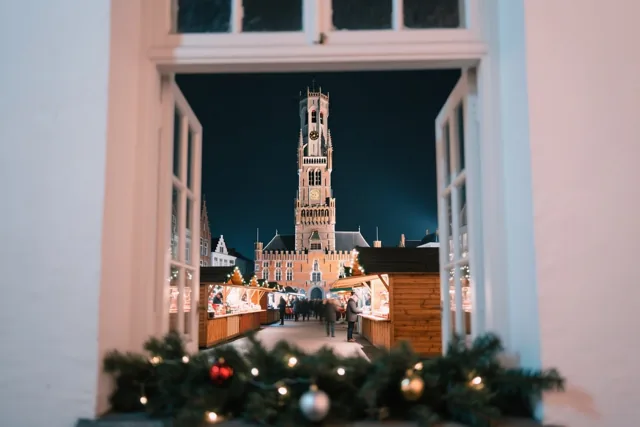 Door een open raam met groen en lichtjes op de vensterbank is een nachtelijke kerstmarkt in Brugge te zien, met houten kraampjes en het verlichte Belfort.