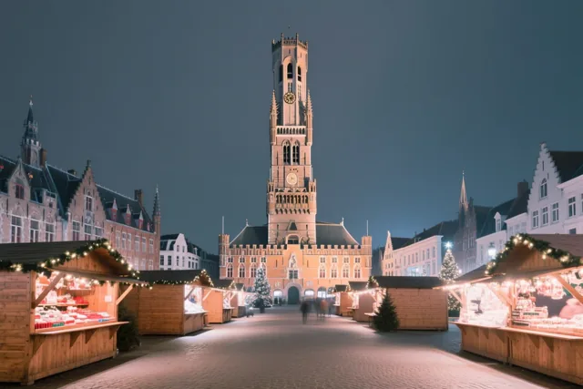 Een Brugse kerstmarkt met houten kraampjes, lichtjes en slingers ligt voor het verlichte Belfort onder een donkerblauwe schemerhemel.