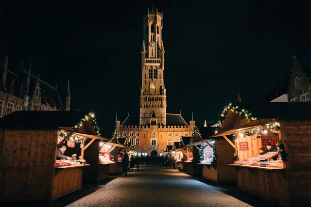 Een nachtelijke kerstmarkt in Brugge heeft houten kraampjes met lichtjes en slingers, met het verlichte Belfort op de achtergrond.