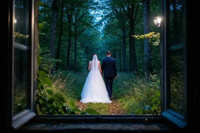 Door een open raam is een bruidspaar van achteren te zien dat wegwandelt over een pad in een dicht groen beukenbos in zacht natuurlijk licht.