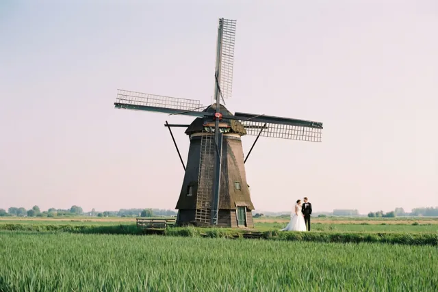 Een bruid en bruidegom staan in de verte in een groen polderveld voor een grote houten windmolen, onder een bleke hemel in zacht licht.