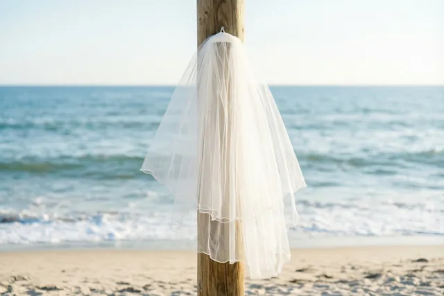 Een witte bruidssluier hangt aan een houten paal op een zandstrand, met de zee en de lucht op de achtergrond in zacht natuurlijk licht.