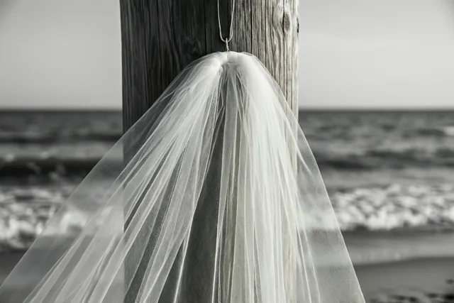 Een zwart-witfoto toont een witte bruidssluier aan een verweerde houten strandpaal, met de zee en golven op de achtergrond.