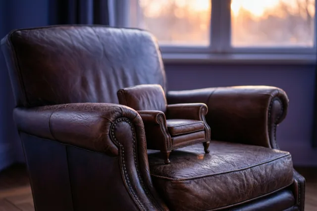 Een grote bruine leren fauteuil en een kleine bijpassende fauteuil staan naast elkaar in een kamer met warm zonlicht door het raam.