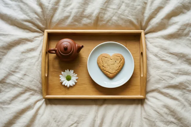 Een houten dienblad met een bruine theepot, een hartvormig koekje op een wit bord en een witte margriet staat op een gekreukt beige dekbed.