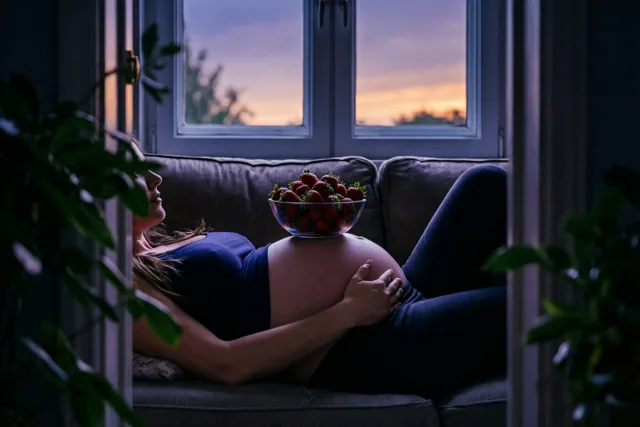 Een zwangere vrouw ligt op een zetel met een schaal aardbeien op haar buik, met een zonsondergang door het raam en diepe blauwe en paarse tinten in de kamer.