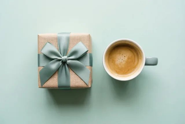 Een vierkant cadeau in beige papier met een blauwgroene strik staat naast een witte mok koffie op een licht blauwgroene ondergrond.