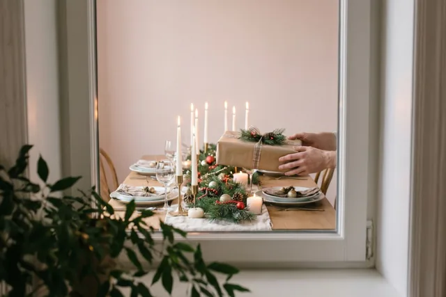 Door een raam zijn handen te zien die een ingepakt kerstcadeau op een feestelijk gedekte tafel met kaarsen en groen leggen, in zachte roze en beige tinten.