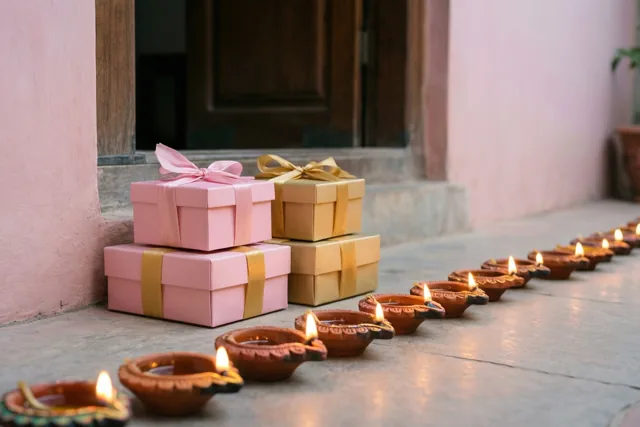 Een rij brandende lampjes van klei staat op een drempel naast stapels roze en gouden cadeaudozen, bij een roze muur en een houten deur.